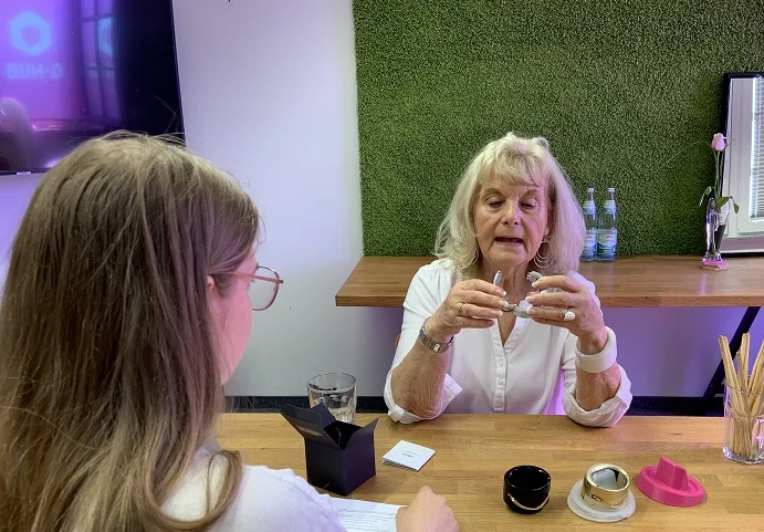 2 Frauen sitzen am Tisch. Eine ist von hinten zu sehen, die andere hält einen Gegenstand in den Händen und schaut sich diesen an