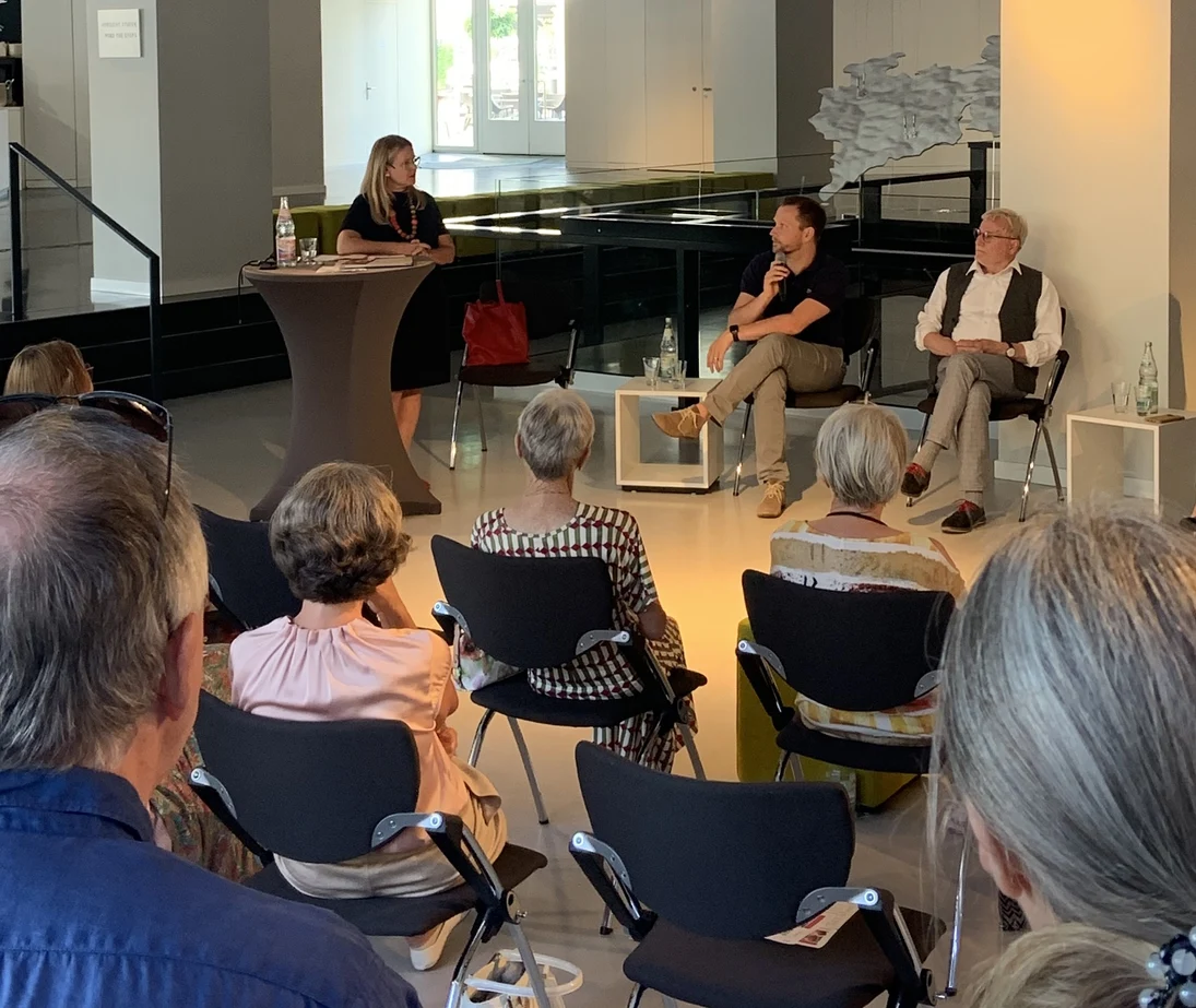 3 Personen sitzen auf einer Bühne und diskutieren vor einem Publikum