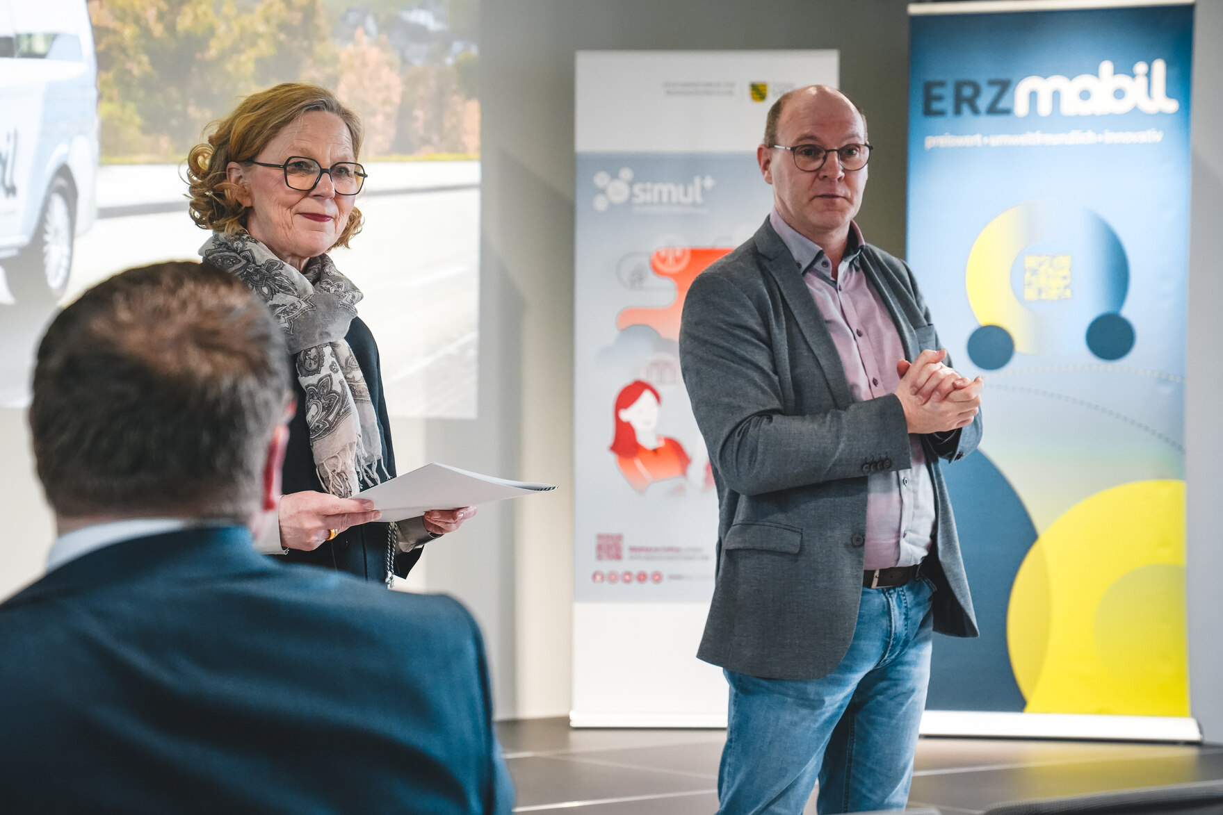 Staatsministerin Kraushaar und der Bürgermeister von Zwönitz Wolfgang Triebert stehen in einem Raum und schauen ins Publikum. Die Ministerin hält eine weiße Mappe in der Hand. Im Hintergrund ist eine Leinwand sowie mehrere Roll-Ups zu sehen.