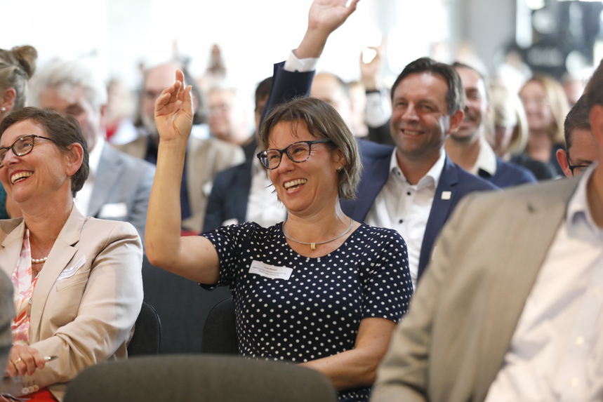 Viele Menschen sitzen auf Stühlen und heben teilweise die Hand und lachen