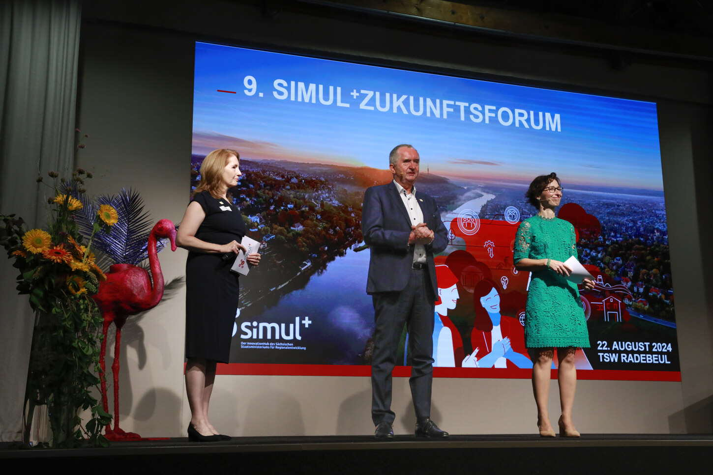 Minister Schmidt steht mit Kristin Hendinger und einer Frau auf der Bühne vor einer Leinwand.